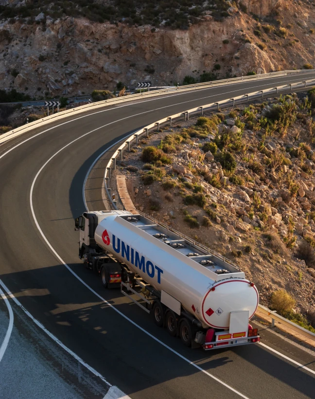 Ciężarówka transportująca ładunek na autostradzie z asfaltowymi pasami i zbiornikiem paliwa. Logo UNIMOT widoczne na pojeździe.